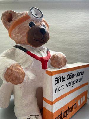 Eine Pappmaché-Figur eines Teddybären in einem weißen Arztkittel. Der Bär trägt einen Kopfspiegel und ein rotes Stethoskop und hält ein Schild hoch, auf dem steht: Bitte Chip-Karte nicht vergessen.