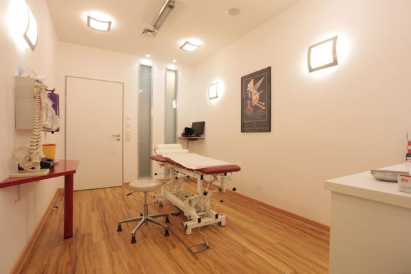 Ein Behandlungsraum mit Holzboden und einer modernen, höhenverstellbaren Therapieliege. Auf einem Beistelltisch steht ein anatomisches Modell der menschlichen Wirbelsäule.