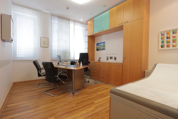 Ein freundliches und helles Sprechzimmer mit einem Schreibtisch, Stühlen für das Arztgespräch und großen Fenstern. An der Seite stehen eine Untersuchungsliege und moderne Holzschränke.